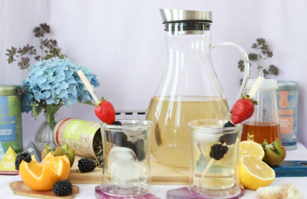 A glass tea pitcher with two cups and fresh berries on a table with fruit as an example of one of the best tea sets for iced tea.