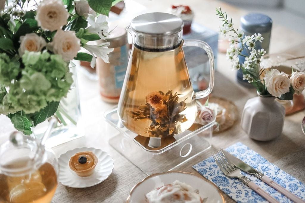 A glass tea maker with a filtering lid holding a blooming tea flower surrounded by fresh flowers and tea pastries.