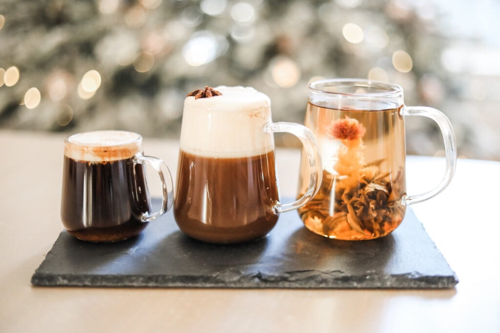 Three tea mugs on a black stone tablet filled with coffee, a chai latte, and floral blooming tea.