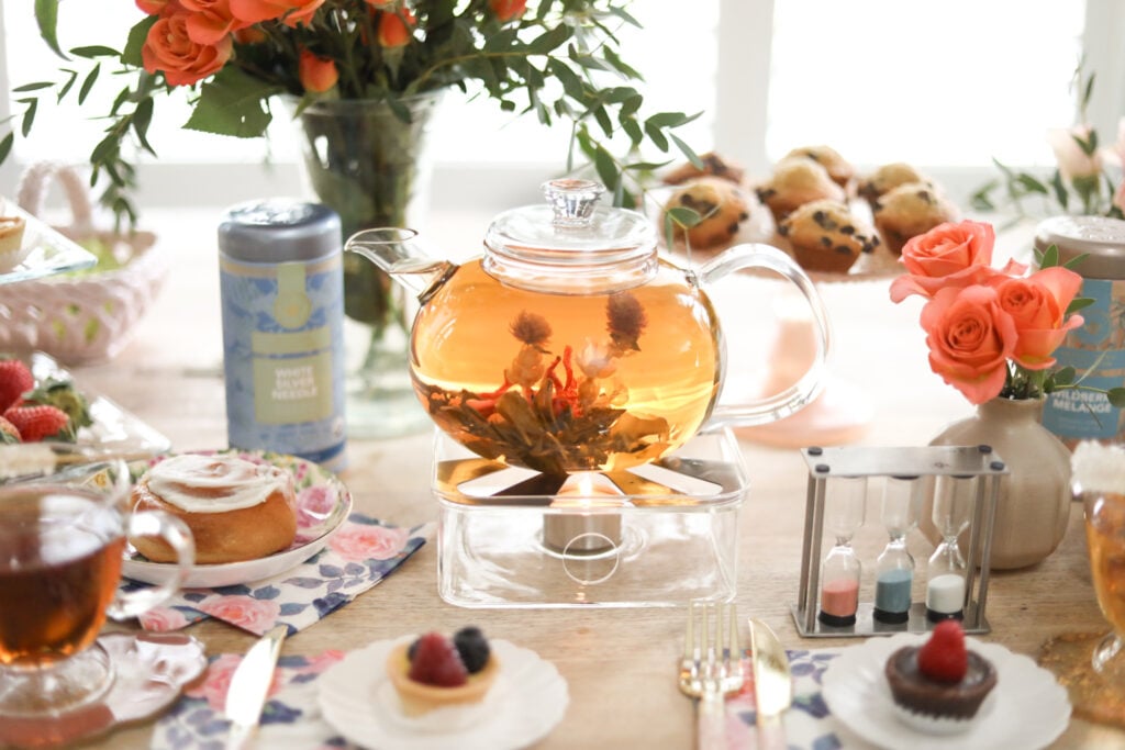 A beautiful glass teapot on a glass warmer surrounded by flowers and accessories and pastries on a table.