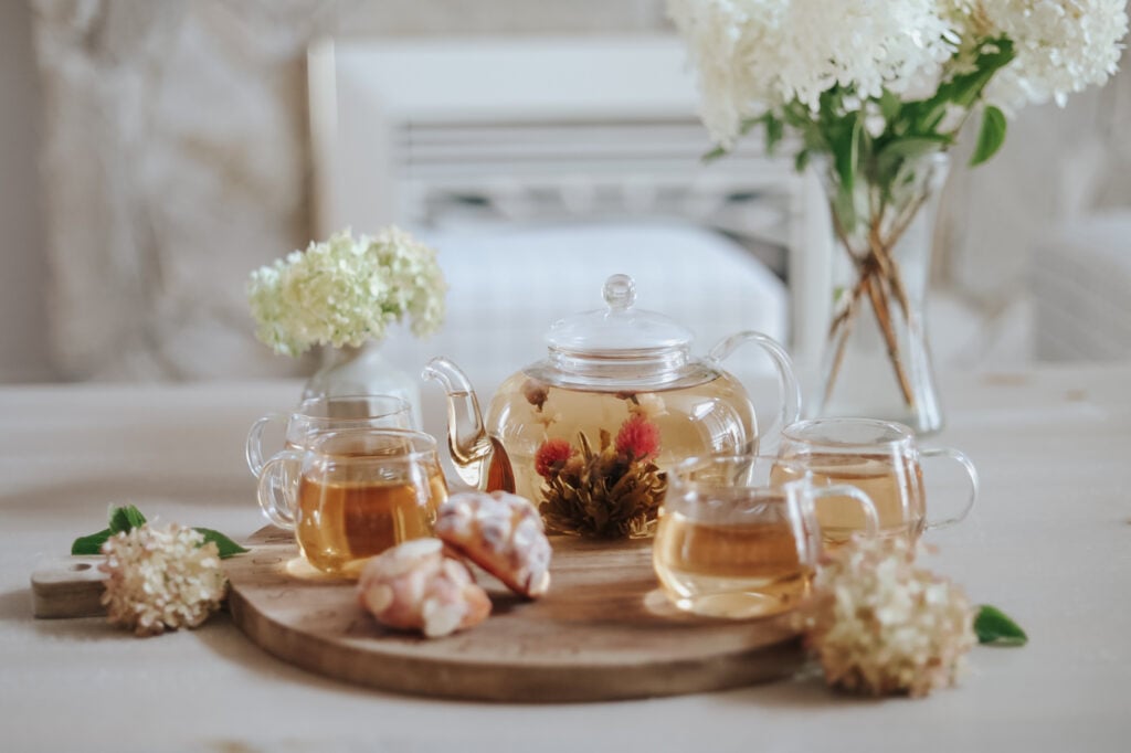 Blossoms glass teapot and teacup set on a wooden display with white flowers and tea pastries.