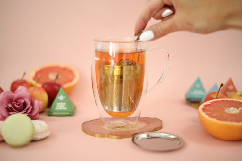 A large glass tea mug with a stainless steel tea infuser on a pink background surrounded by tea sachets, flowers, and citrus slices.