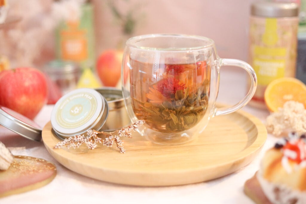 A large glass tea mug with a blooming flower tea on a wooden stand with a loose-leaf tea canister, flowers, and pastries.