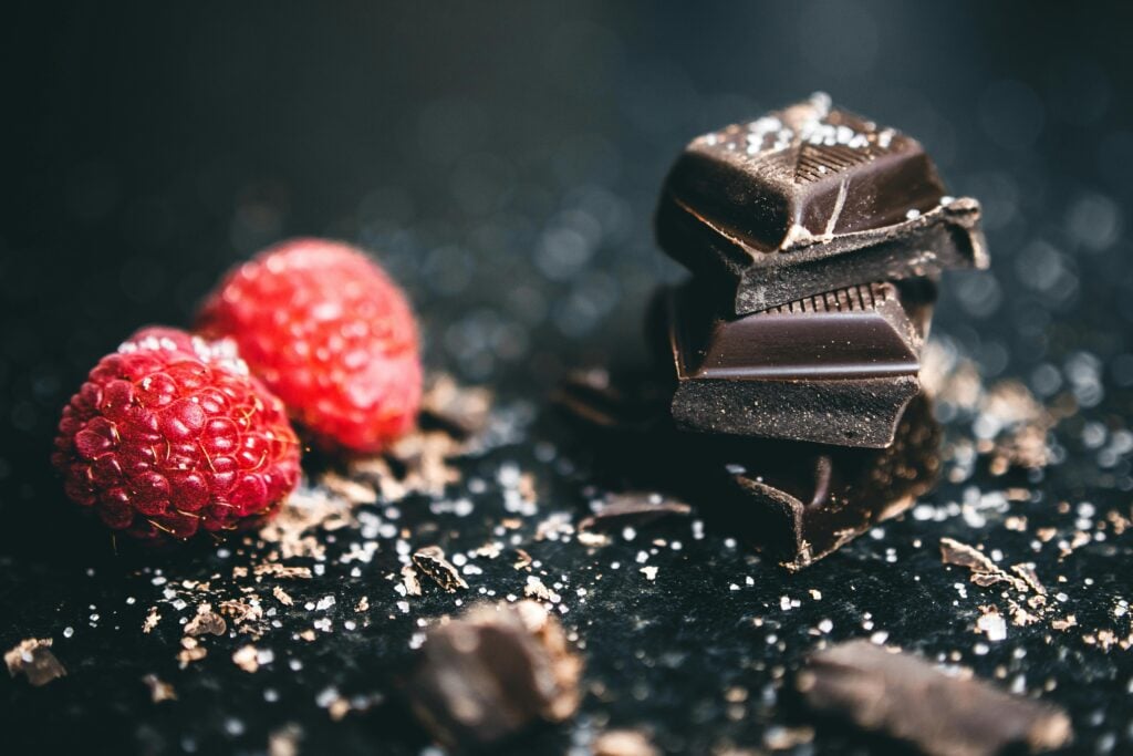 Fresh raspberries and chocolate squares stacked on a black table with a sprinkle of sugar and salt.
