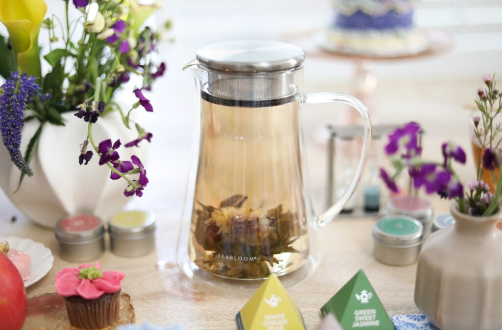 A tall, modern glass teapot with blooming flower tea on a table with fresh flowers, pastries, and tea sachets.