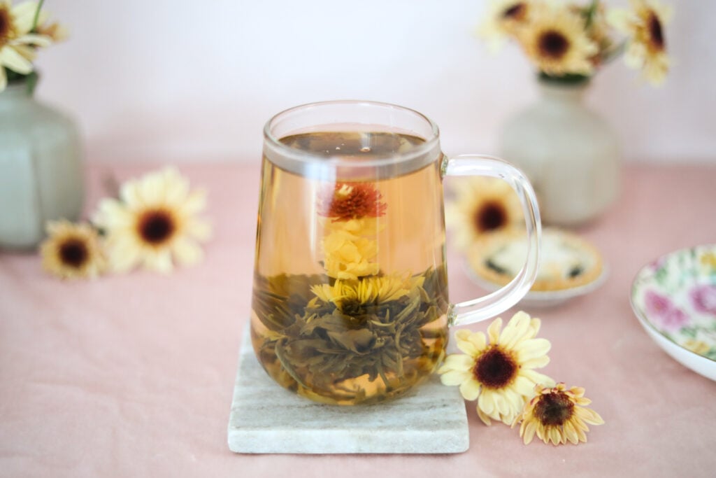 A tapered glass tea mug with a blooming flower tea on a marble coaster surrounded by fresh daisies.
