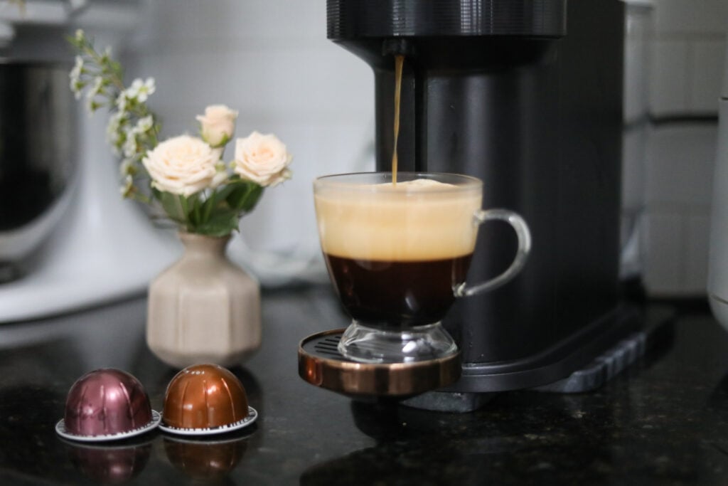 A glass teacup with coffee on an espresso maker with two espresso pods and a vase of white flowers.