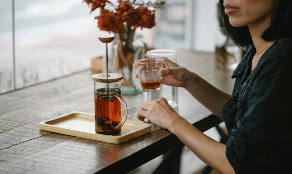 A woman holding a glass double-walled teacup with a tea press on a wooden table.