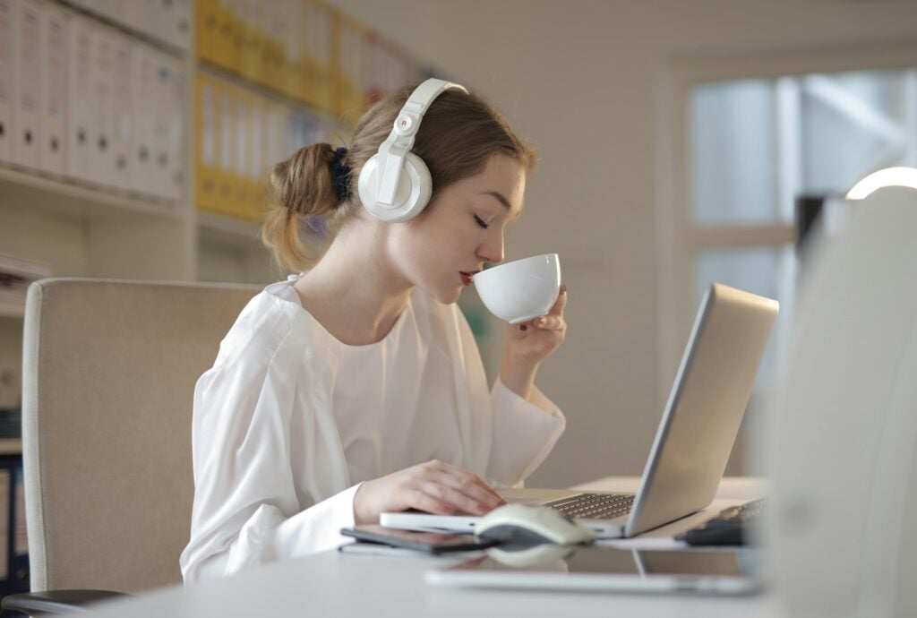A young woman dressed in white sitting at a table with her laptop while listening to music and sipping on tea for anxiety.